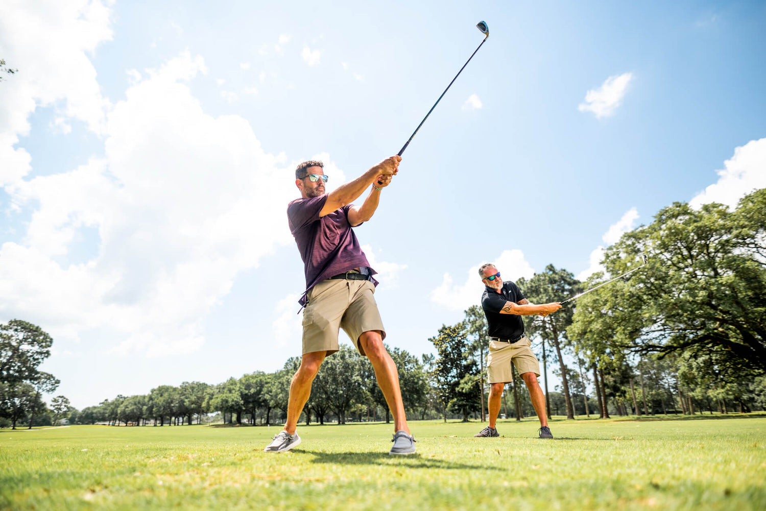Two men on golf course swinging clubs, each wearing MSC Stretch Fit Shorts in Oyster and dark polos